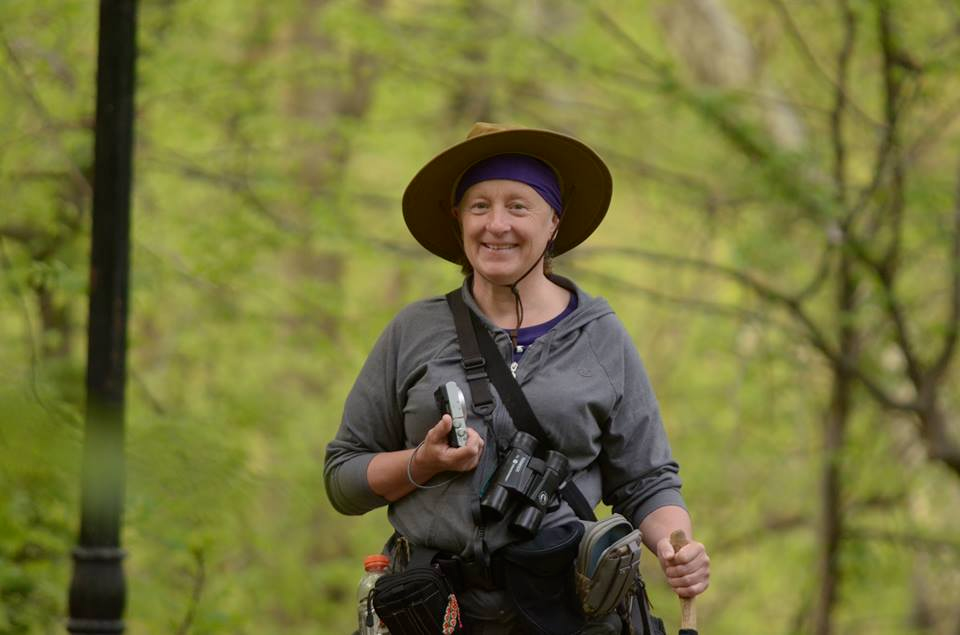 Lee smiling in a wide-brimmed hat with binoculars in Central Park in spring